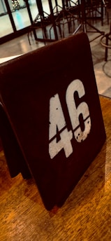 A rustic wooden table number sign displaying the number 46 in bold, white numerals. The background consists of a polished wooden table surface and bar stools in a restaurant or cafe setting. Glass doors and a hint of decoration are visible in the background, suggesting an indoor venue.
