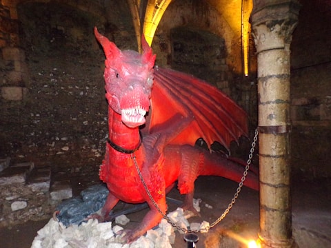 A large red dragon sculpture is situated in a dimly lit, cave-like setting. The dragon is adorned with a chain around its neck and is posed with an open mouth displaying sharp teeth. The background features stone walls and pillars, illuminated by a warm yellow light.