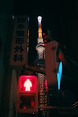 A brightly lit tower stands against a dark night sky, its structure illuminated in vivid colors. In the foreground, a red pedestrian traffic light signals to stop, while adjacent signage features Japanese characters.