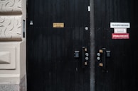 A dark wooden door with multiple signs attached to it, including one indicating a sauna and others related to fire safety and a sprinkler system. The door has visible locks and number pads. It is part of a building with textured beige walls.