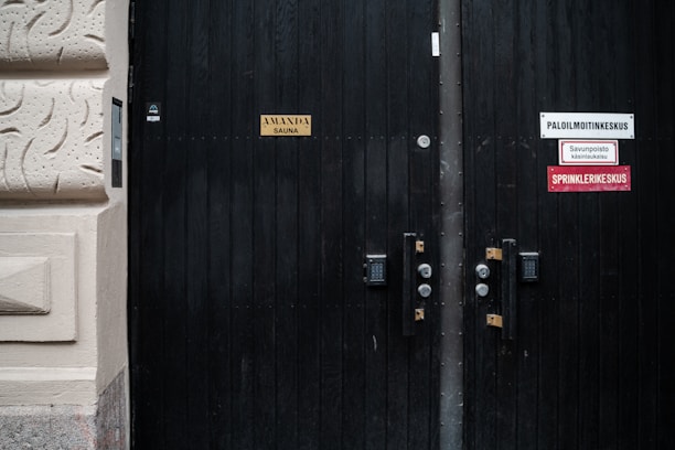 A dark wooden door with multiple signs attached to it, including one indicating a sauna and others related to fire safety and a sprinkler system. The door has visible locks and number pads. It is part of a building with textured beige walls.