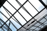 A geometric framework of steel beams and glass panes forms a large, intricate pattern against the sky. The structure creates a crisscrossed grid, with individual sections allowing light to pass through.