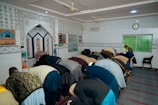 A group of people are engaging in prayer within a decorated interior space, featuring Arabic calligraphy and colorful geometric patterns on the walls. The individuals are in a bowing position, aligned in several rows on a striped carpet. The room has a few chairs, a clock, and a digital display indicating prayer times.