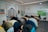 A group of people are engaging in prayer within a decorated interior space, featuring Arabic calligraphy and colorful geometric patterns on the walls. The individuals are in a bowing position, aligned in several rows on a striped carpet. The room has a few chairs, a clock, and a digital display indicating prayer times.