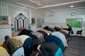 A group of people are engaging in prayer within a decorated interior space, featuring Arabic calligraphy and colorful geometric patterns on the walls. The individuals are in a bowing position, aligned in several rows on a striped carpet. The room has a few chairs, a clock, and a digital display indicating prayer times.
