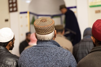 A group of men is gathered, most wearing intricately designed caps. They are facing away, suggesting participation in a communal or religious activity. The setting appears to be indoors, possibly a place of worship.