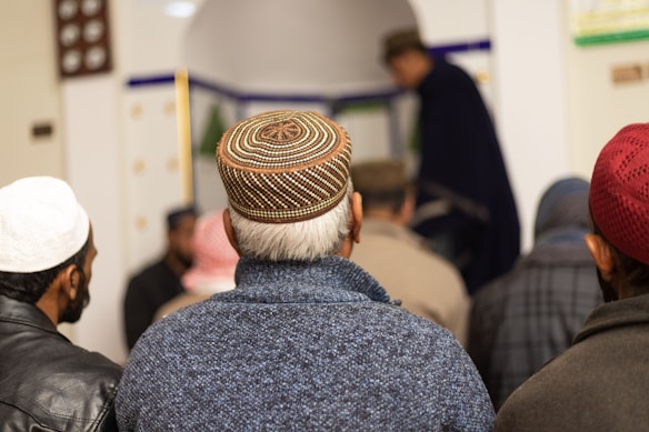 A group of men is gathered, most wearing intricately designed caps. They are facing away, suggesting participation in a communal or religious activity. The setting appears to be indoors, possibly a place of worship.