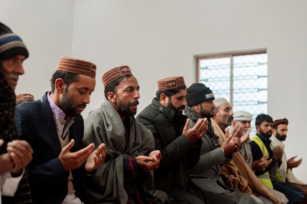 A group of men seated in a row, engaging in prayer with their hands raised. They are dressed warmly with a combination of jackets and traditional caps, suggesting a cultural or religious gathering. The setting appears to be indoors with a window in the background.