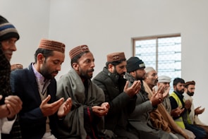 A group of men seated in a row, engaging in prayer with their hands raised. They are dressed warmly with a combination of jackets and traditional caps, suggesting a cultural or religious gathering. The setting appears to be indoors with a window in the background.