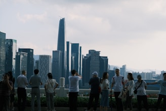 A group of people stands on a viewing platform overlooking a modern city skyline with tall skyscrapers against a partly cloudy sky.