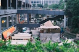 A bustling urban area features a variety of shops and outdoor stalls with a modern architectural design. People walk along a pathway lined with greenery on the sides. Above, a walkway connects parts of the building. There is a large tent-like structure visible in the center, possibly part of a market or outdoor event.