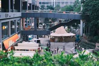 A bustling urban area features a variety of shops and outdoor stalls with a modern architectural design. People walk along a pathway lined with greenery on the sides. Above, a walkway connects parts of the building. There is a large tent-like structure visible in the center, possibly part of a market or outdoor event.