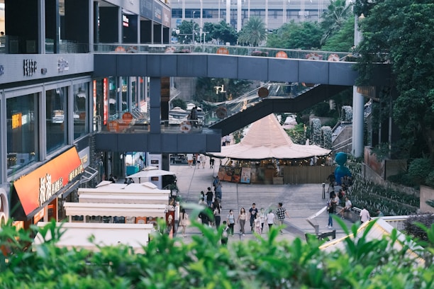 A bustling urban area features a variety of shops and outdoor stalls with a modern architectural design. People walk along a pathway lined with greenery on the sides. Above, a walkway connects parts of the building. There is a large tent-like structure visible in the center, possibly part of a market or outdoor event.