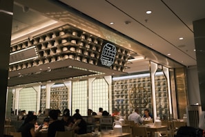 The interior of a modern restaurant with several people dining at wooden tables. The design features shelves with decorative items and soft lighting, creating a cozy and inviting atmosphere. The ceiling displays geometric patterns and there are illuminated signs with decorative elements on the walls.