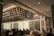 The interior of a modern restaurant with several people dining at wooden tables. The design features shelves with decorative items and soft lighting, creating a cozy and inviting atmosphere. The ceiling displays geometric patterns and there are illuminated signs with decorative elements on the walls.