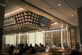 The interior of a modern restaurant with several people dining at wooden tables. The design features shelves with decorative items and soft lighting, creating a cozy and inviting atmosphere. The ceiling displays geometric patterns and there are illuminated signs with decorative elements on the walls.