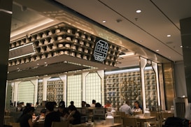 The interior of a modern restaurant with several people dining at wooden tables. The design features shelves with decorative items and soft lighting, creating a cozy and inviting atmosphere. The ceiling displays geometric patterns and there are illuminated signs with decorative elements on the walls.