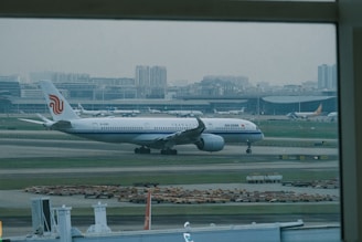 An airplane with the Air China livery is taxiing on the runway of a large airport, with the terminal buildings and city skyline in the background. Several other aircraft are parked near the terminal.