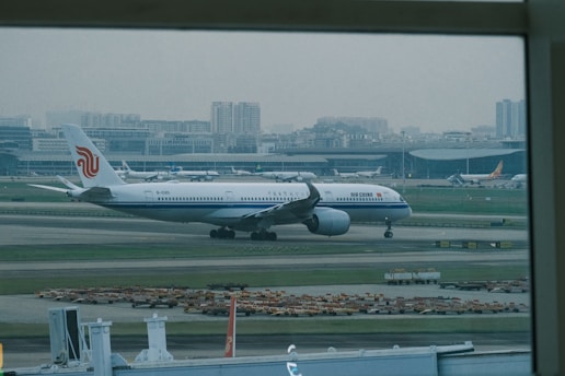 An airplane with the Air China livery is taxiing on the runway of a large airport, with the terminal buildings and city skyline in the background. Several other aircraft are parked near the terminal.