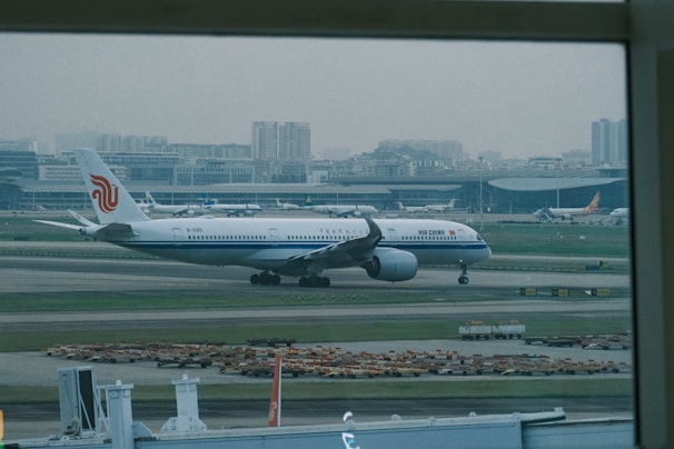 An airplane with the Air China livery is taxiing on the runway of a large airport, with the terminal buildings and city skyline in the background. Several other aircraft are parked near the terminal.