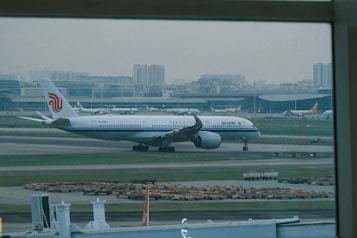 An airplane with the Air China livery is taxiing on the runway of a large airport, with the terminal buildings and city skyline in the background. Several other aircraft are parked near the terminal.
