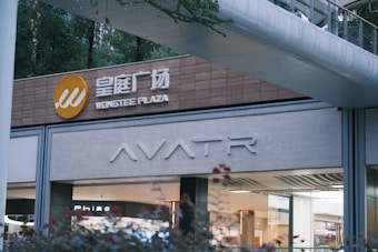 A modern shopping plaza storefront with signage for Wongtee Plaza in both English and Chinese. The design features clean lines and neutral tones. A walkway arches overhead, and greenery is visible in the background.