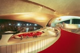 A futuristic, expansive interior featuring sweeping curves and modern architectural elements. The space is defined by a large, open area with a red carpeted walkway, wide ceilings, and an array of flags in the distance. There is sleek furniture in the center with people gathered around.