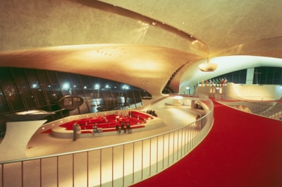 A futuristic, expansive interior featuring sweeping curves and modern architectural elements. The space is defined by a large, open area with a red carpeted walkway, wide ceilings, and an array of flags in the distance. There is sleek furniture in the center with people gathered around.