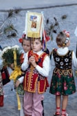 Children are dressed in vibrant traditional costumes, carrying peacock feathers and flowers. They hold a colorful banner with images. The street setting adds context to the cultural event.