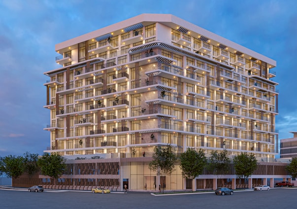 A modern multi-story residential building with a sleek design. The structure features numerous balconies and large windows, creating a pattern on its facade. The building is lit up, indicating a dusk or evening scene. Several vehicles are parked or driving on the street, and there are trees planted along the sidewalk.