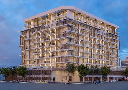 A modern multi-story residential building with a sleek design. The structure features numerous balconies and large windows, creating a pattern on its facade. The building is lit up, indicating a dusk or evening scene. Several vehicles are parked or driving on the street, and there are trees planted along the sidewalk.