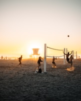 A group of people playing beach volleyball at sunset, with additional people walking and standing around. A lifeguard tower is visible in the background, positioned directly in front of the setting sun, creating a silhouette effect.