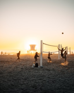 A group of people playing beach volleyball at sunset, with additional people walking and standing around. A lifeguard tower is visible in the background, positioned directly in front of the setting sun, creating a silhouette effect.