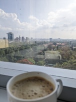 A steaming cup of coffee beside a window overlooking green coffee plantations.