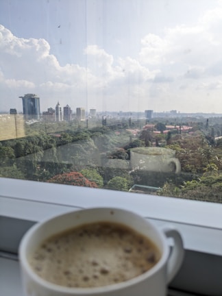 A steaming cup of hot coffee with swirling cream, placed beside a window overlooking lush green forest.