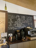 A cozy cafe interior featuring a chalkboard with artistic drawings and witty coffee-related quotes. The board includes a drawing of a person sipping coffee, phrases like 'Are you a chai-vinist??' and 'I can't espresso what you really bean to me!!'. The countertop displays various objects including a kettle, a large water dispenser, coffee cups, and other coffee-making equipment. A decorative green bottle hangs from the ceiling.
