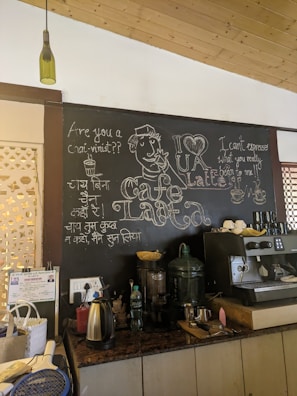 A cozy cafe interior featuring a chalkboard with artistic drawings and witty coffee-related quotes. The board includes a drawing of a person sipping coffee, phrases like 'Are you a chai-vinist??' and 'I can't espresso what you really bean to me!!'. The countertop displays various objects including a kettle, a large water dispenser, coffee cups, and other coffee-making equipment. A decorative green bottle hangs from the ceiling.