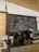A cozy cafe interior featuring a chalkboard with artistic drawings and witty coffee-related quotes. The board includes a drawing of a person sipping coffee, phrases like 'Are you a chai-vinist??' and 'I can't espresso what you really bean to me!!'. The countertop displays various objects including a kettle, a large water dispenser, coffee cups, and other coffee-making equipment. A decorative green bottle hangs from the ceiling.