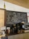 A cozy cafe interior featuring a chalkboard with artistic drawings and witty coffee-related quotes. The board includes a drawing of a person sipping coffee, phrases like 'Are you a chai-vinist??' and 'I can't espresso what you really bean to me!!'. The countertop displays various objects including a kettle, a large water dispenser, coffee cups, and other coffee-making equipment. A decorative green bottle hangs from the ceiling.