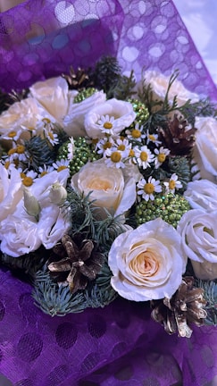 A beautifully arranged floral bouquet featuring white roses, small daisy-like flowers with yellow centers, green foliage, and pinecones. The bouquet is wrapped in a purple, textured paper with a polka dot pattern.