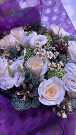 A beautifully arranged floral bouquet featuring white roses, small daisy-like flowers with yellow centers, green foliage, and pinecones. The bouquet is wrapped in a purple, textured paper with a polka dot pattern.