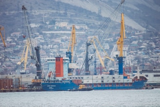 A busy shipyard scene featuring multiple cranes and a large cargo ship. The backdrop consists of a densely built hillside town, creating a bustling industrial atmosphere.