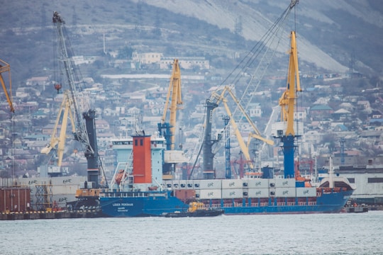 A busy shipyard scene featuring multiple cranes and a large cargo ship. The backdrop consists of a densely built hillside town, creating a bustling industrial atmosphere.