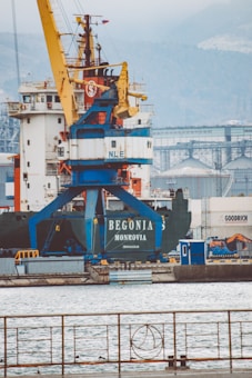 A large cargo ship with the name 'Begonia Monrovia' is docked at an industrial port. A prominent yellow and blue crane is positioned over the vessel, and several shipping containers are visible in the background. The surrounding area includes industrial buildings and structures.