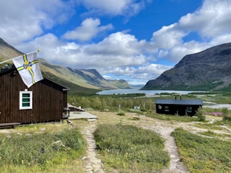 A picturesque landscape featuring two wooden cabins in a lush, green mountainous area. A path of stone slabs winds through the grass towards the buildings. A flag with a yellow and blue pattern flutters beside one cabin. There is a wide river or lake in the background, framed by expansive mountains and a sky filled with scattered clouds.