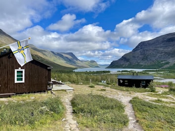 A picturesque landscape featuring two wooden cabins in a lush, green mountainous area. A path of stone slabs winds through the grass towards the buildings. A flag with a yellow and blue pattern flutters beside one cabin. There is a wide river or lake in the background, framed by expansive mountains and a sky filled with scattered clouds.