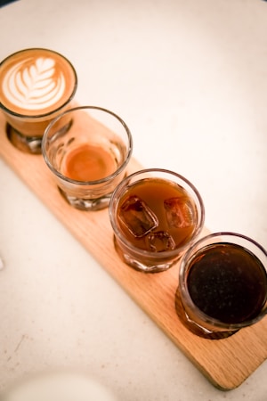 Four glasses of coffee are arranged in a row on a wooden tray. The first glass contains a latte with a delicate leaf pattern in the foam. The second glass has a small amount of espresso. The third glass holds an iced coffee with visible ice cubes. The last glass contains a dark coffee, possibly cold brew.