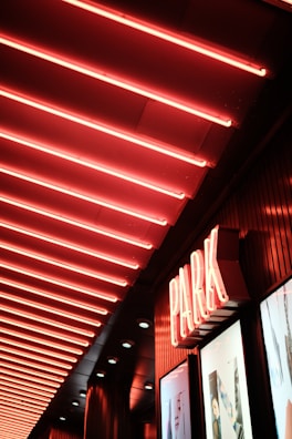 A neon-lit ceiling features a series of parallel red lights creating a vibrant and dynamic atmosphere. Below the ceiling, a sign reading 'PARK' in neon lights is prominently displayed above a storefront with photographic advertisements.