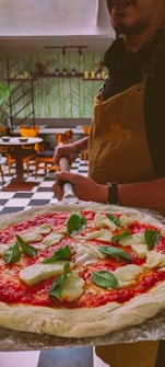 A person in a brown apron is holding a pizza peel with a freshly prepared Margherita pizza, topped with slices of mozzarella and fresh basil leaves. The background shows a cozy dining area with green patterned wallpaper, wooden chairs, and neatly arranged shelves.