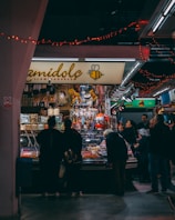 A bustling market scene with a group of people gathered around a stall selling a variety of colorful candies and goods. The stall is decorated with festive red string lights, and the name of the shop is displayed prominently with a bee logo. The background is filled with more stalls and bright lighting, indicating an indoor setting.